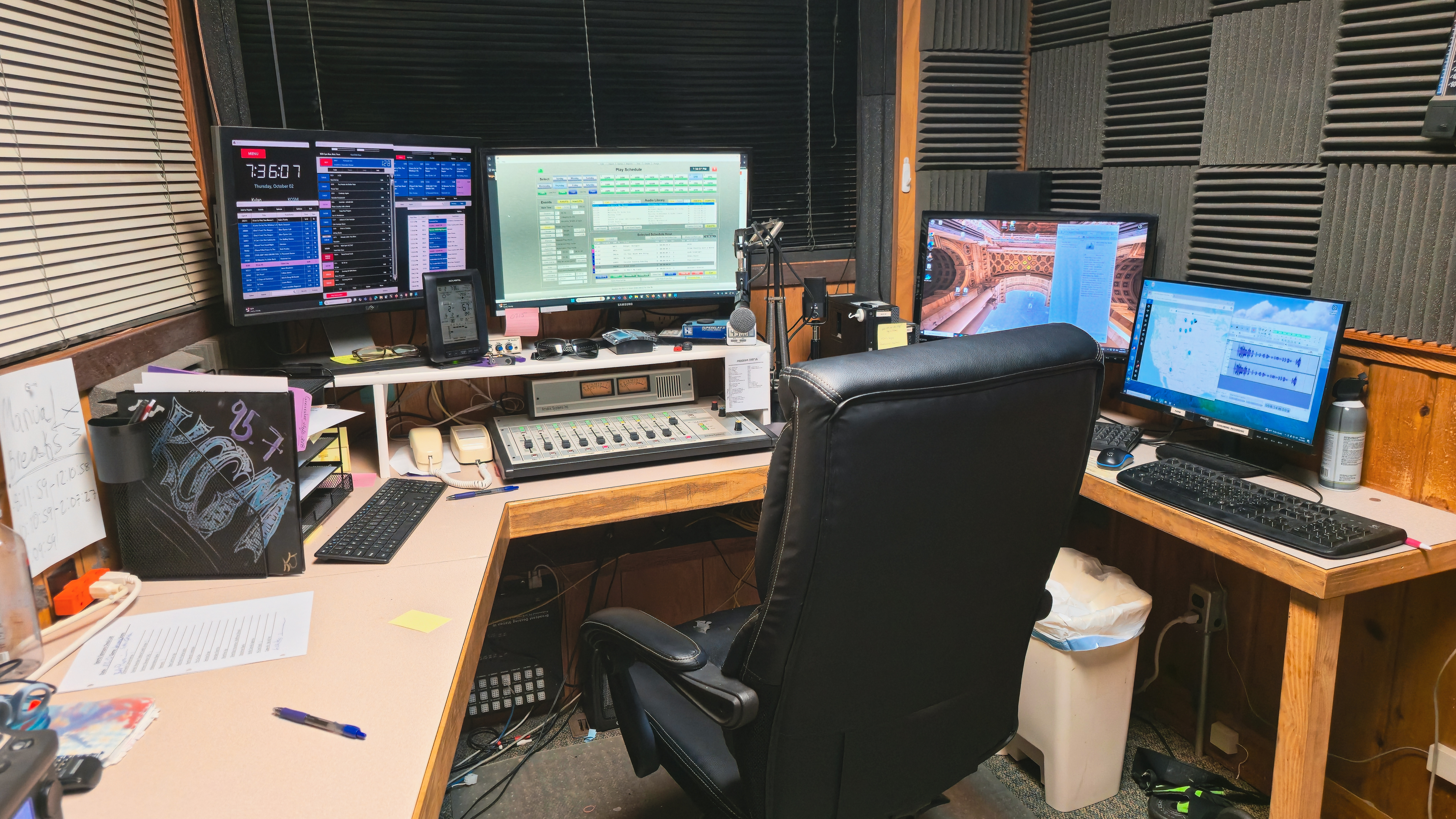 A radio broadcasting studio with multiple computer monitors displaying software, a sound mixing console, and a comfortable office chair.