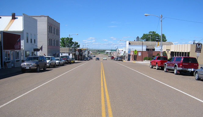 View of a small town main street lined with parked cars and buildings on both sides under a clear blue sky.